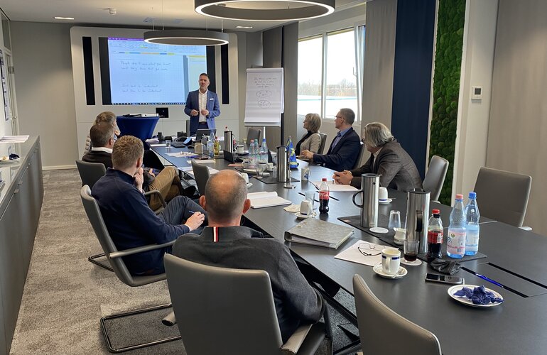 Mehrere Leute sitzen an einem langen Tisch und einer hält eine Präsentation.
