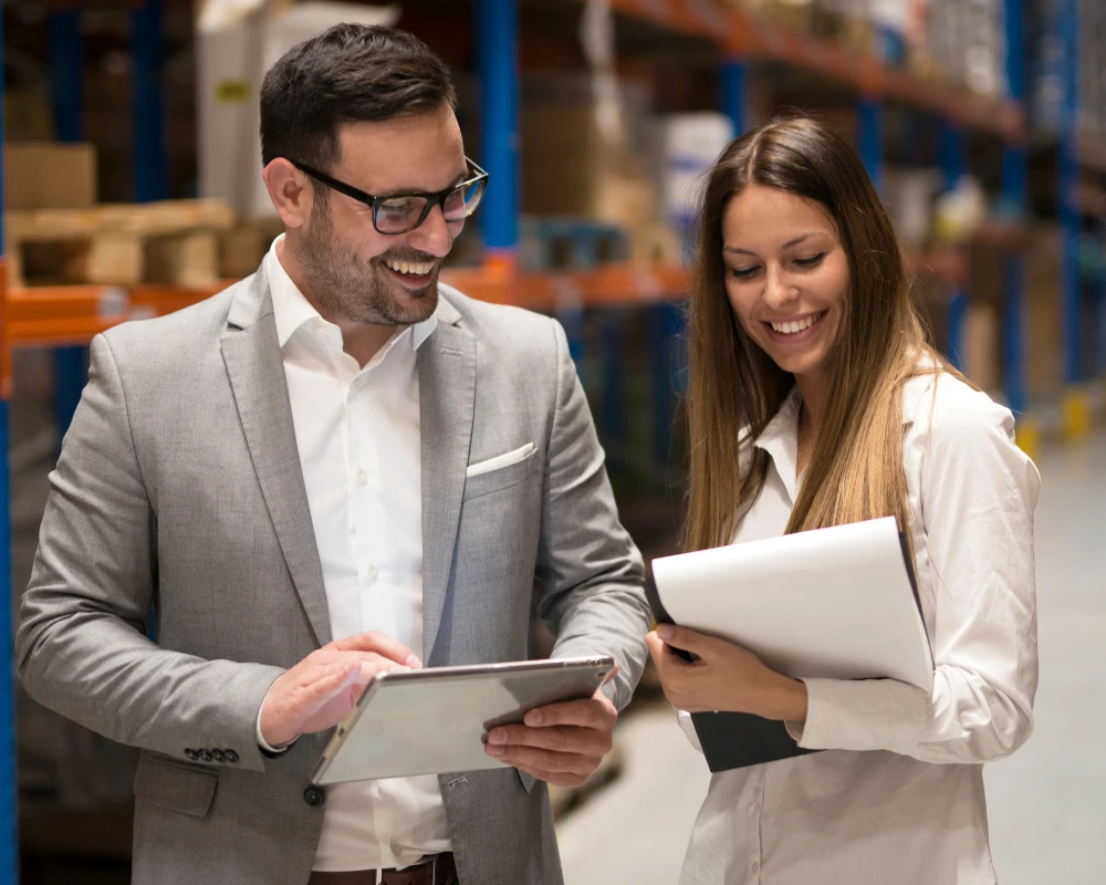 Zwei Personen im Lagerhaus, die gemeinsam auf ein Tablet und Dokumente schauen und lächeln.