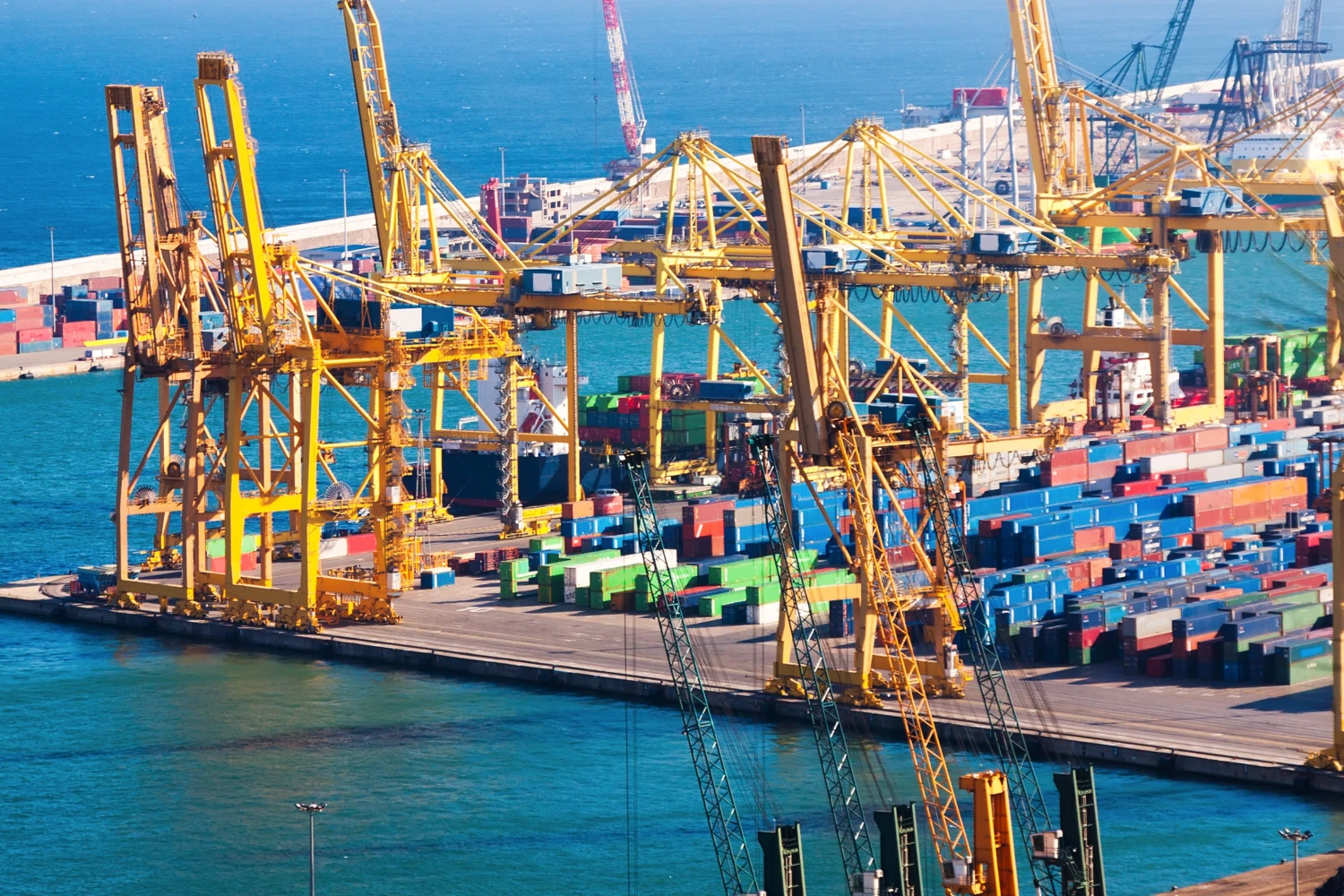 Port with container ships, numerous colorful container stacks and large yellow harbor cranes on the water.