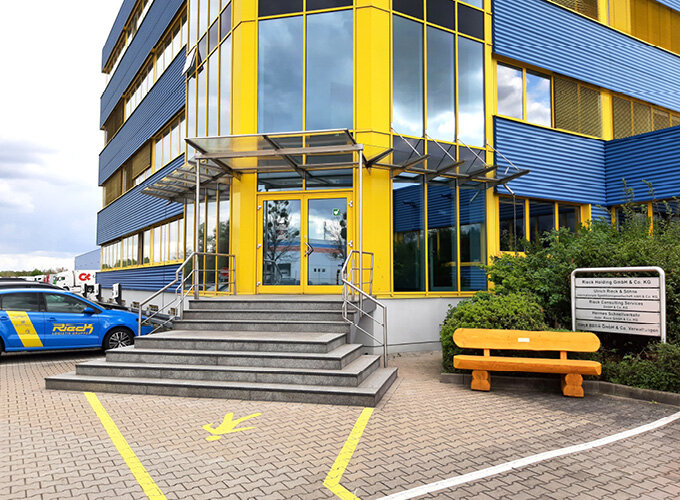 Eingang eines modernen Bürogebäudes mit gelben und blauen Fassadenelementen, Treppe, gelber Bank und parkendem blauen Auto.