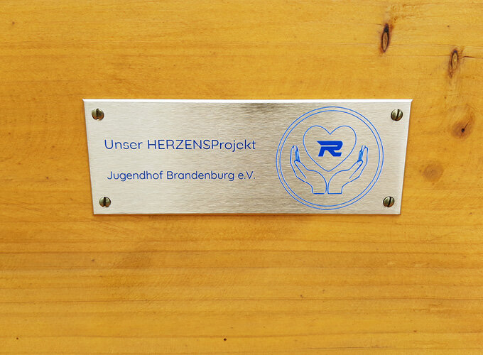 Metallschild mit der Aufschrift Unser HERZENSPROjekt Jugendhof Brandenburg e.V. auf Holz befestigt.