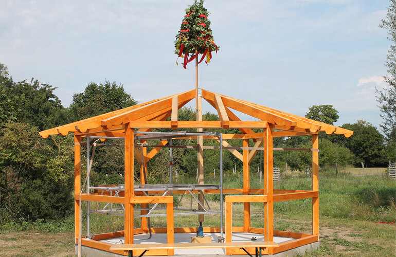 Rohbau eines Holzhauses mit Richtkranz auf dem Dach, umgeben von Wiese und Bäumen.