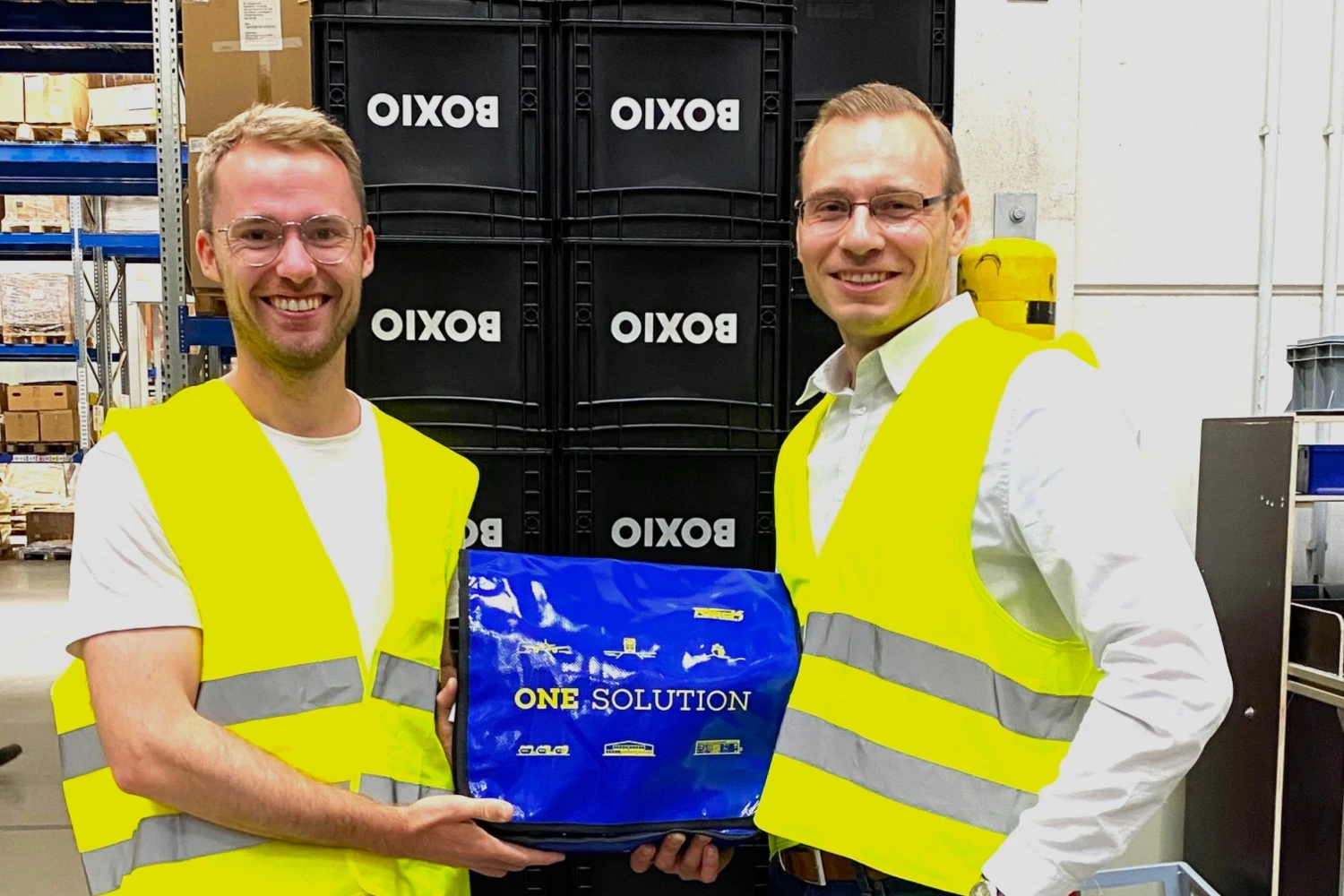 Zwei Männer mit gelben Warnwesten halten gemeinsam eine blaue Box in einem Lagerraum.
