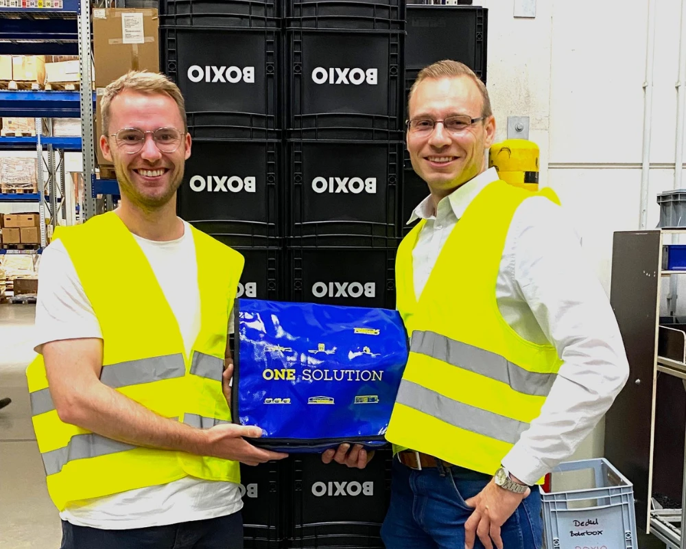 Zwei Männer mit gelben Sicherheitswesten halten eine blaue Box in einem Lagerraum.