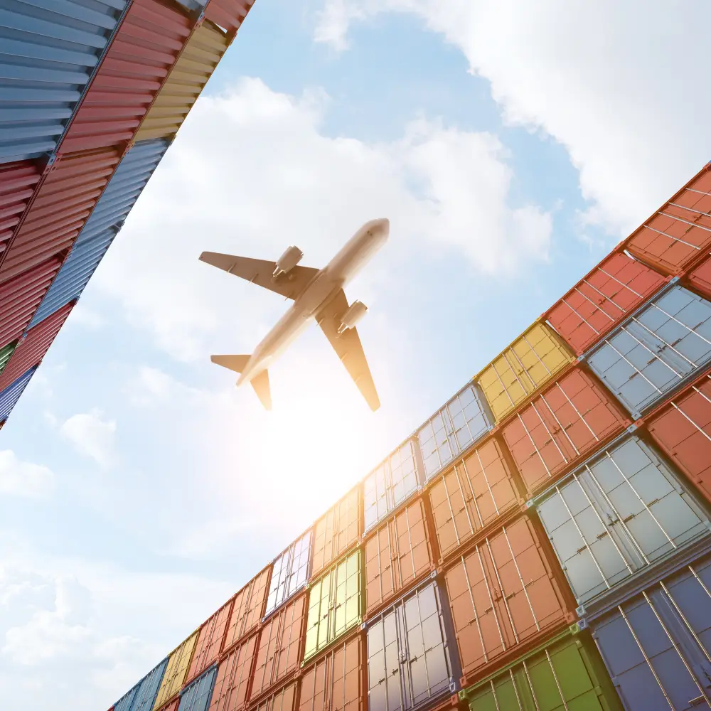 An airplane flies over colorful cargo containers at the port in the sunshine.