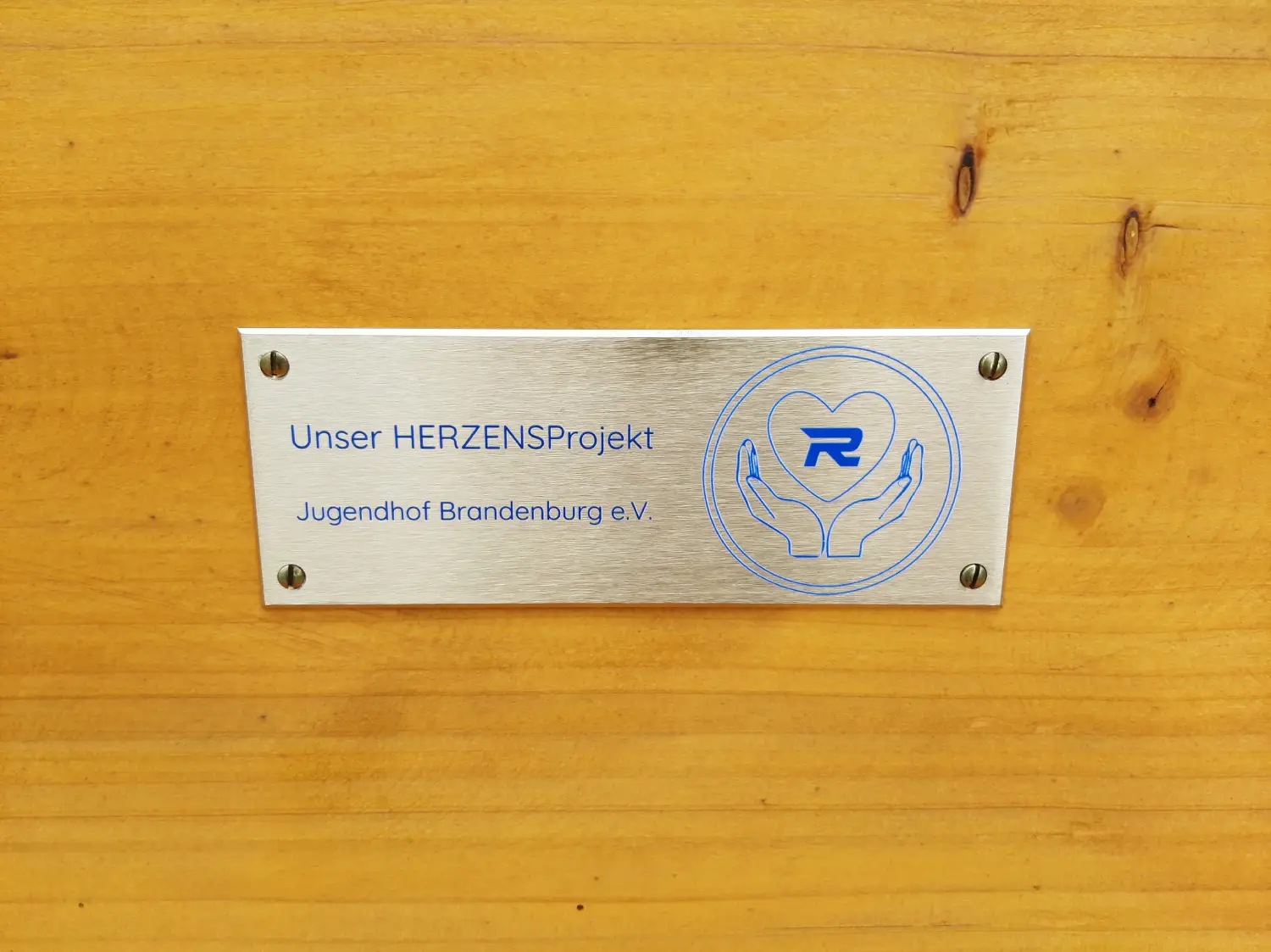 Metallschild mit der Aufschrift "Unser HERZENSProjekt Jugendhof Brandenburg e.V." und einem Logo mit zwei Händen.