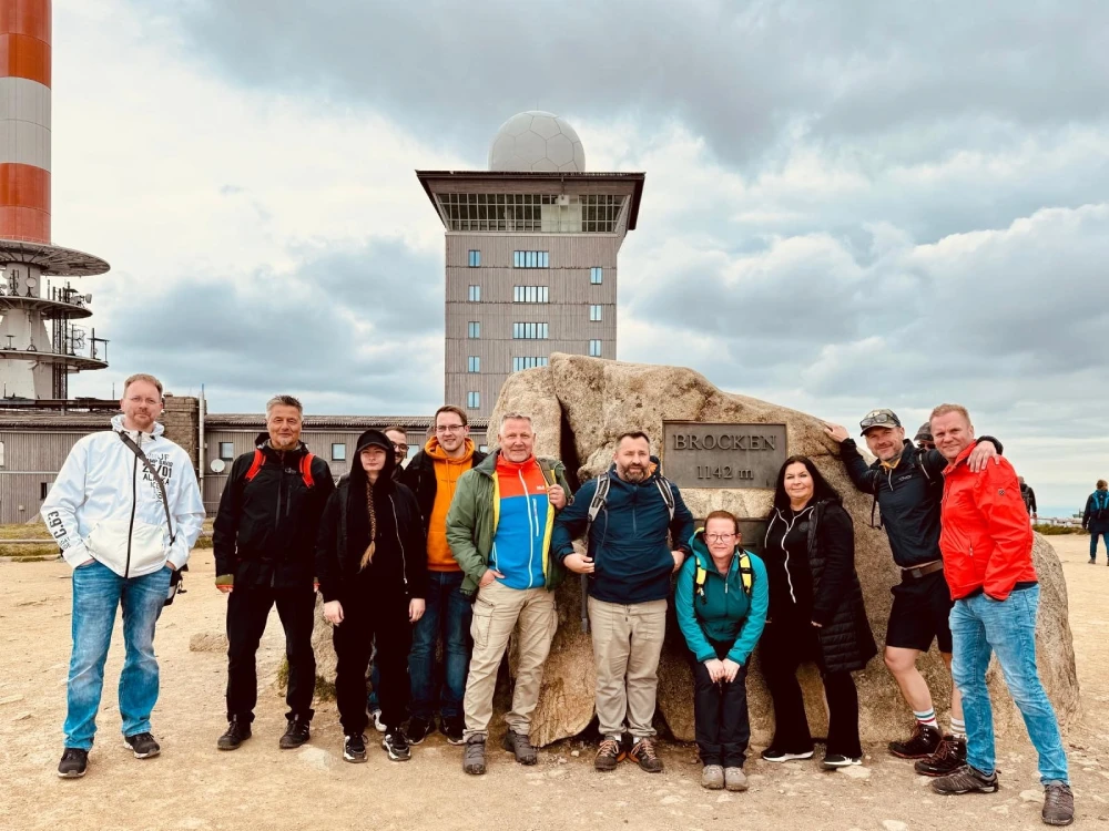 Gruppe von zehn Personen steht vor einem großen Stein mit der Aufschrift "Brocken" unter bewölktem Himmel.