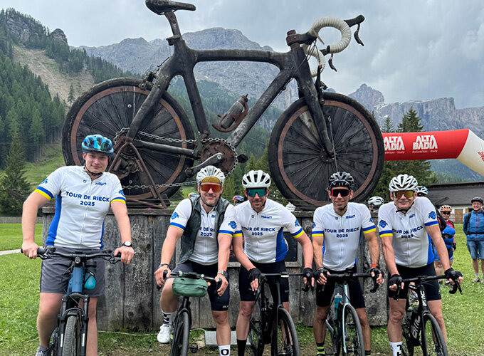 Fünf Radfahrer in Rennradkleidung posieren vor einer großen Fahrradskulptur aus Holz in einer Berglandschaft.