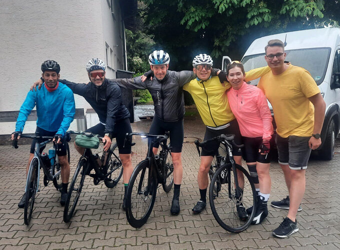 Fünf Radfahrer in Sportkleidung posieren mit ihren Fahrrädern, daneben steht eine Person in gelbem T-Shirt und Shorts, im Hintergrund ein weißer Transporter.