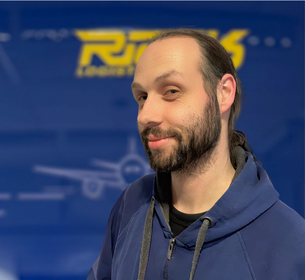 Porträt eines Mannes mit Bart und langem Haar, der eine blaue Kapuzenjacke trägt, vor einem unscharfen blauen Hintergrund mit gelbem Schriftzug.