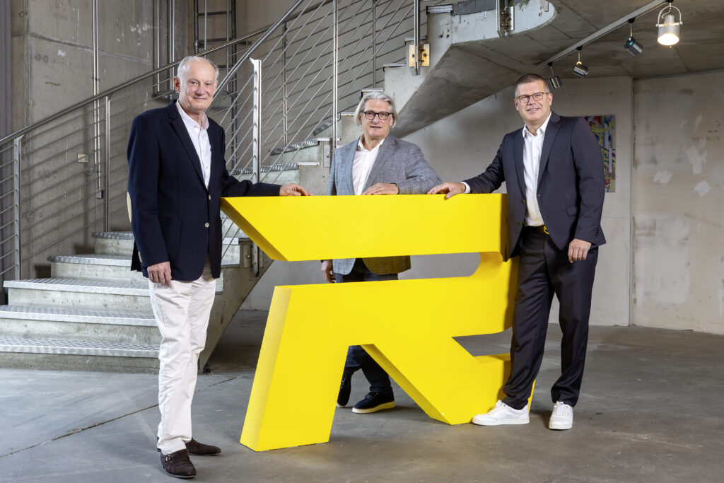 Three men in suits stand around a large yellow symbol in a modern interior with a staircase.